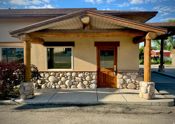 Rustic-inspired exterior featuring a wood-finish door with transom window, stone veneer siding, and natural log columns supporting a pitched roofline