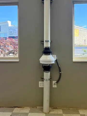 Wall-mounted white PVC piping with an integrated exhaust fan and electrical outlets, set against a grey wall with checkered flooring