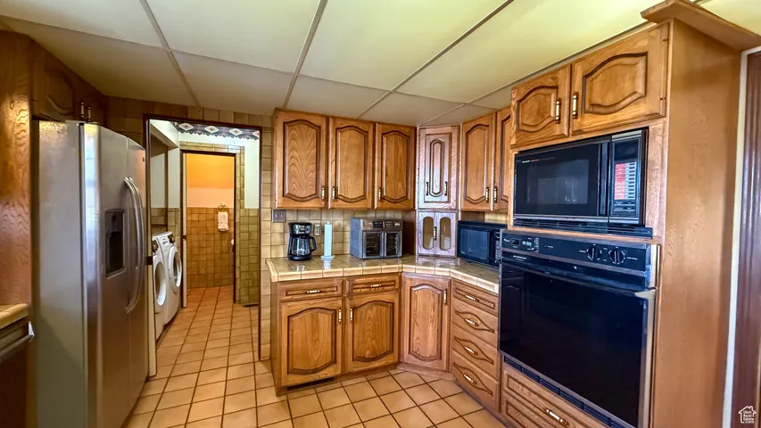 Kitchen with brown cabinetry, light tile patterned floors, black appliances, a drop ceiling, and tile counters