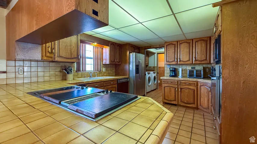 Kitchen featuring brown cabinets, a drop ceiling, tile countertops, tasteful backsplash, and washing machine and clothes dryer