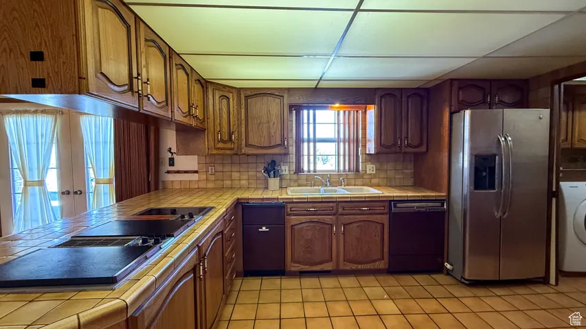 Kitchen with tile countertops, black appliances, brown cabinets, washer / dryer, and tasteful backsplash