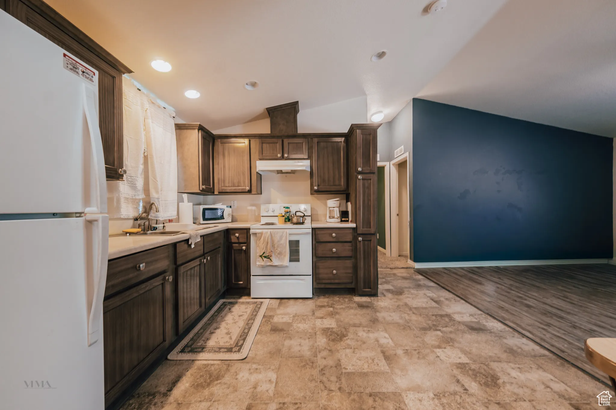 Kitchen featuring white appliances, light countertops, lofted ceiling, dark brown cabinetry, and under cabinet range hood