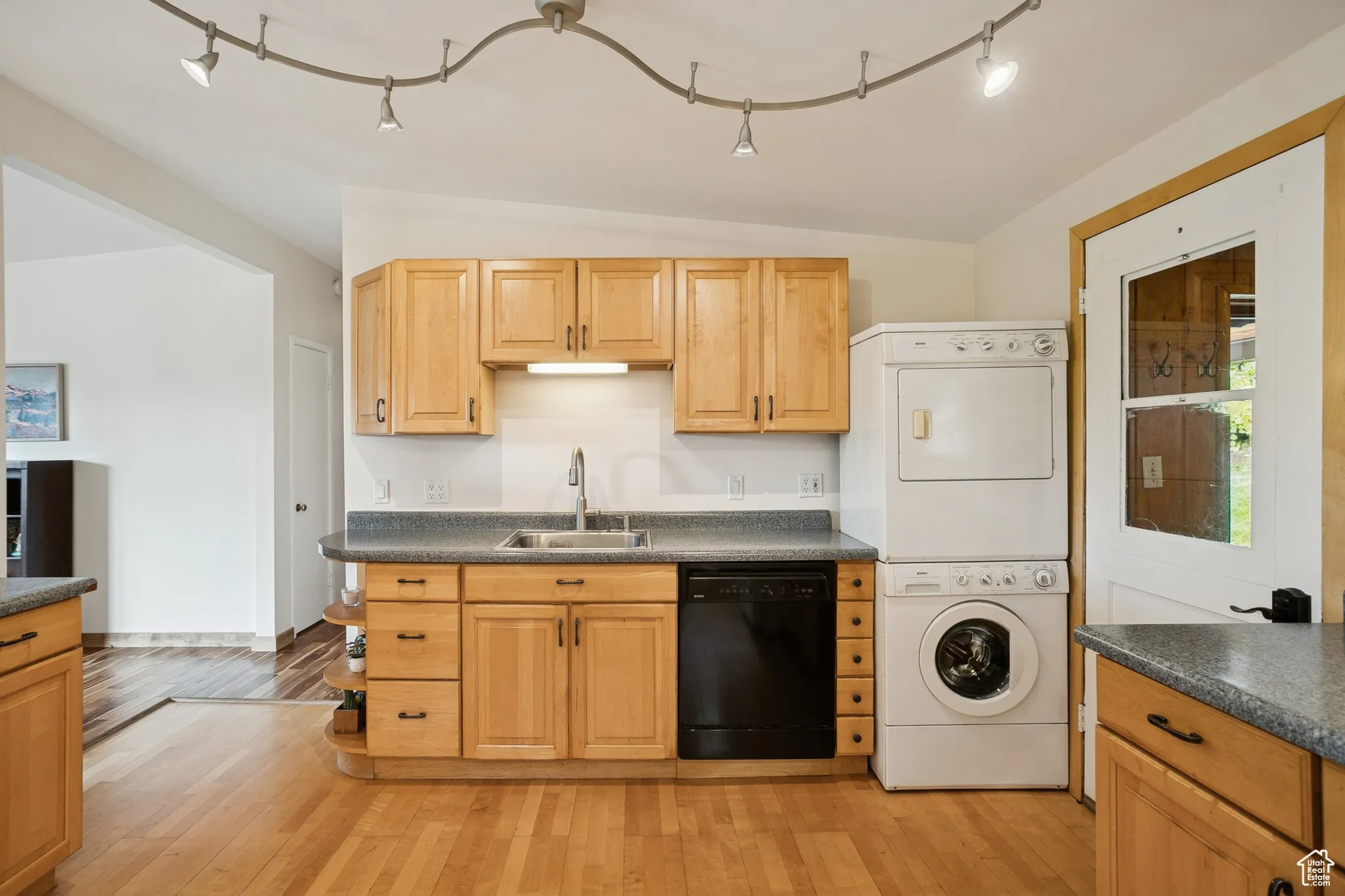 Kitchen with rail lighting, dark countertops, light wood finished floors, black dishwasher, and stacked washing machine and dryer