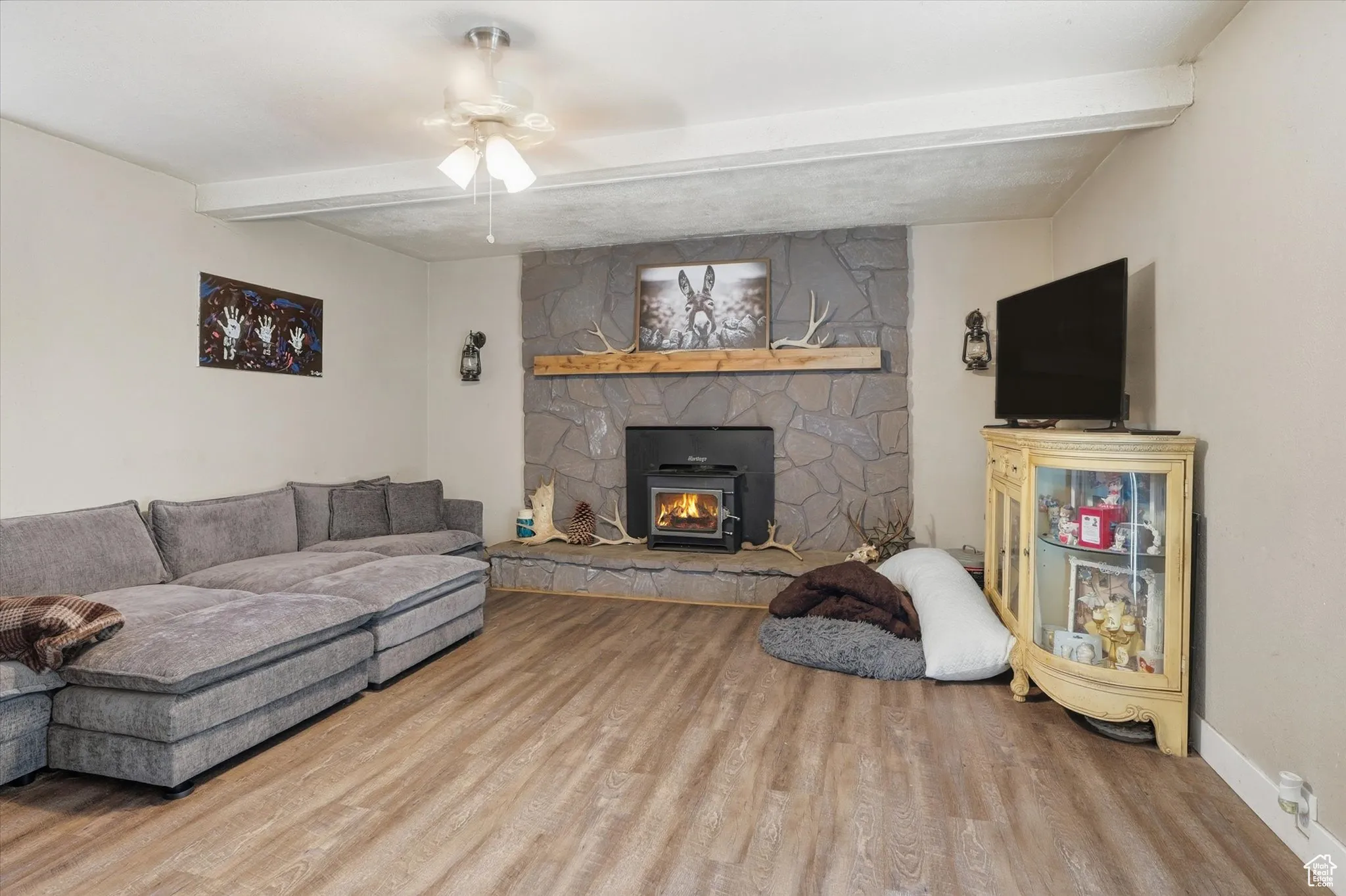 Living area with beamed ceiling, a ceiling fan, wood finished floors, and a wood stove