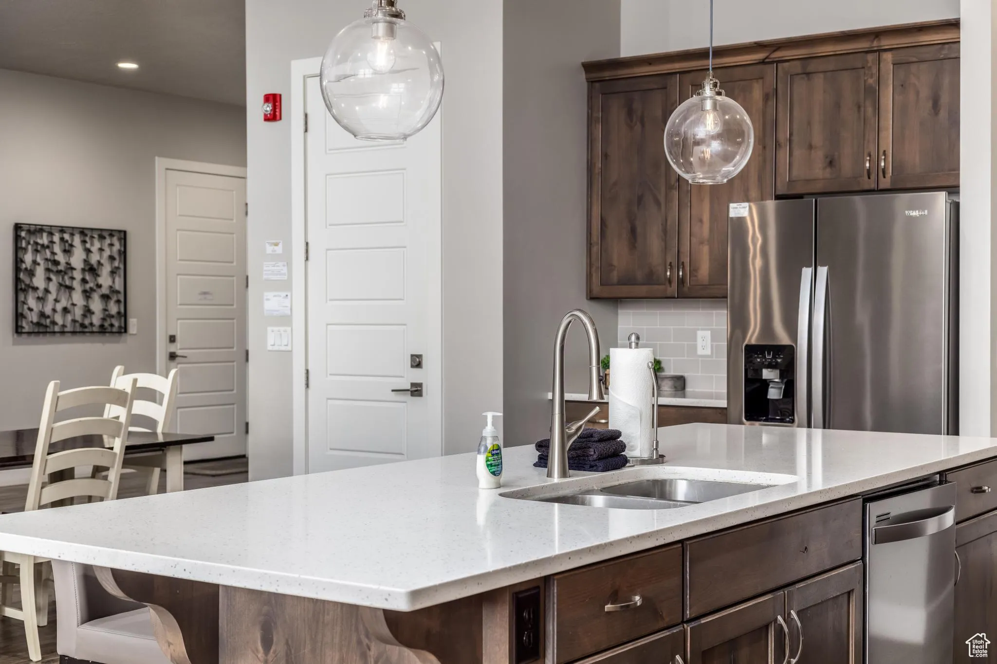 Kitchen with dark brown cabinetry, stainless steel fridge with ice dispenser, tasteful backsplash, light stone counters, and hanging light fixtures