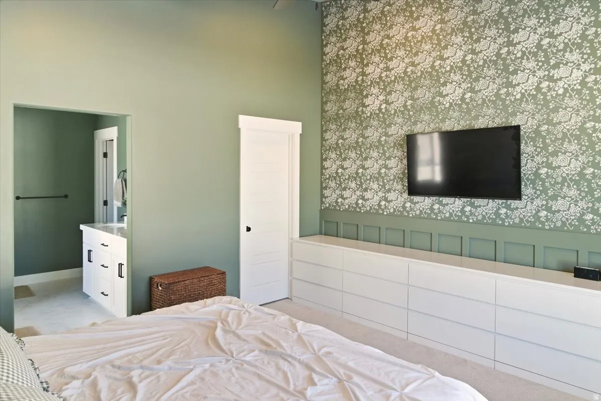 Bedroom with wallpapered walls, a wainscoted wall, light colored carpet, ensuite bathroom, and a high ceiling