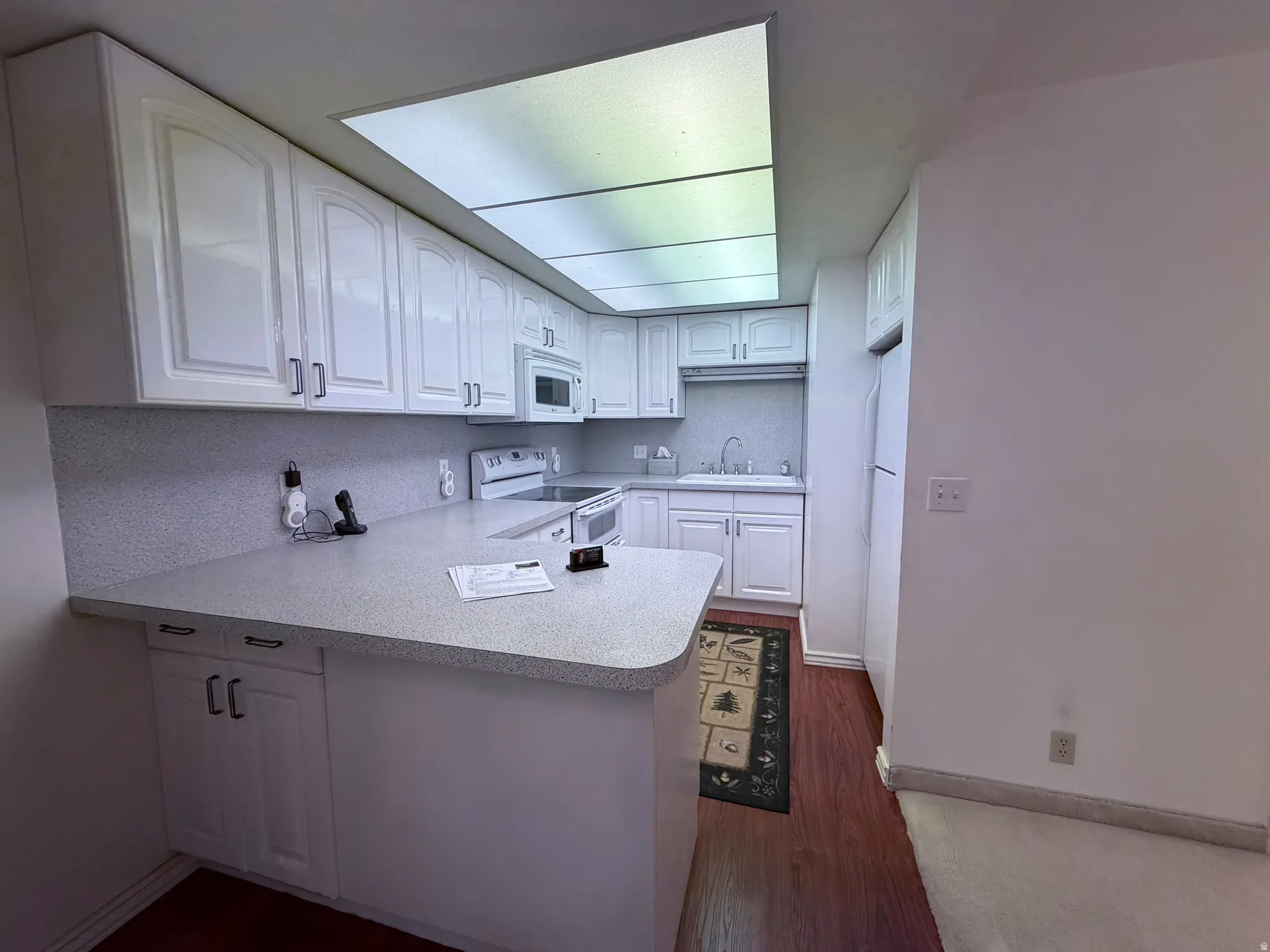 Kitchen featuring white cabinetry, a peninsula, light countertops, white appliances, and dark wood-style flooring