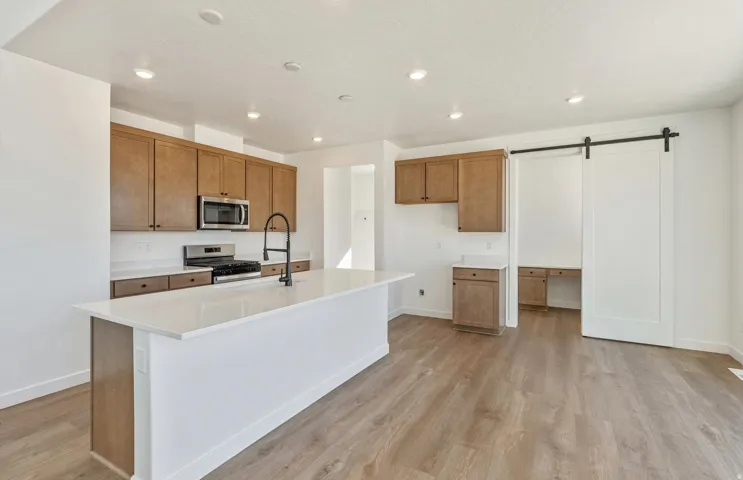 Kitchen with brown cabinetry, a barn door, stainless steel appliances, an island with sink, and light wood-type flooring