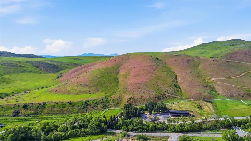 Expansive rural landscape featuring rolling green and brown hills, a dark-roofed building surrounded by trees, and a paved road with white fencing