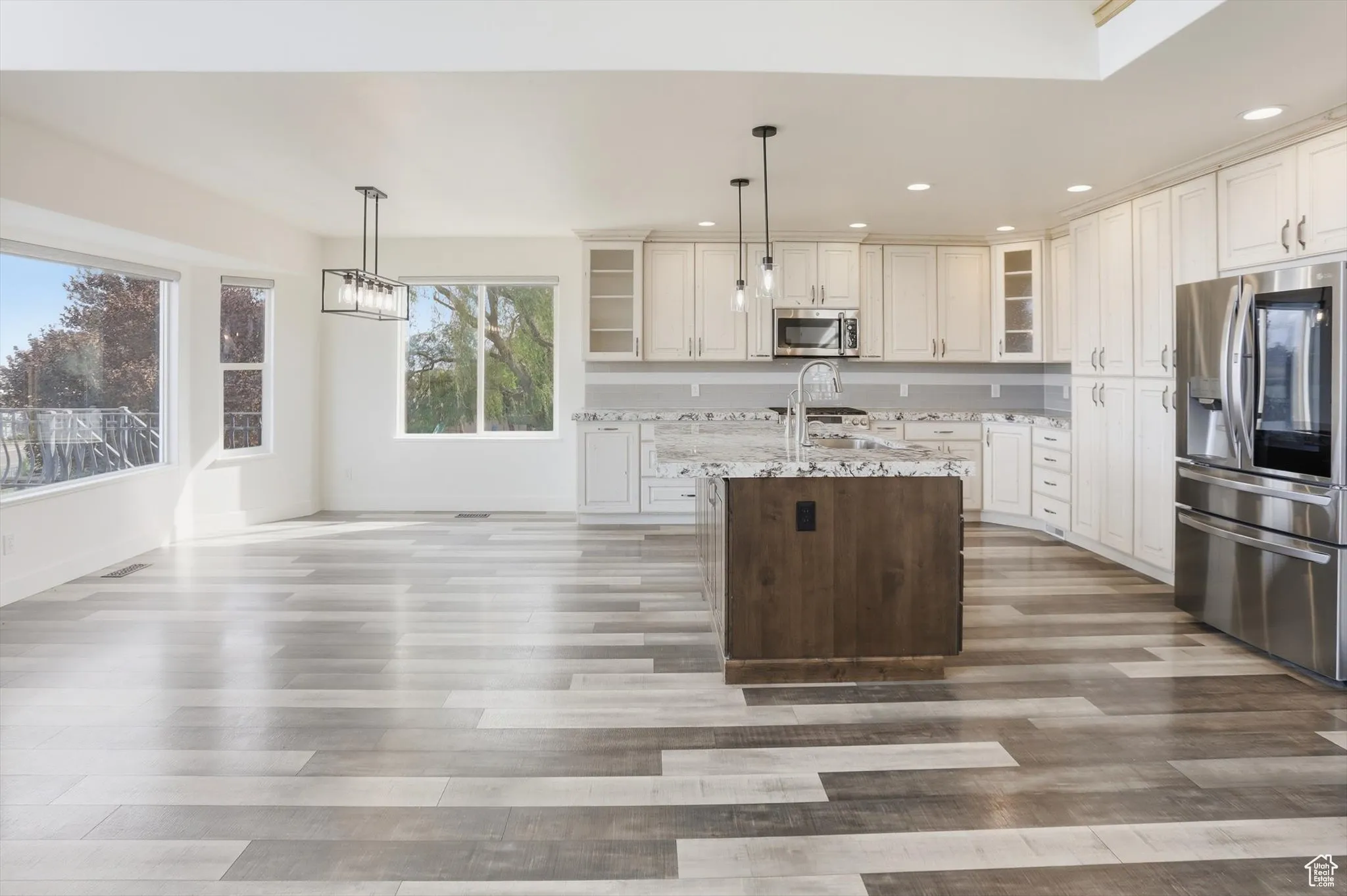 Kitchen with glass insert cabinets, stainless steel appliances, decorative light fixtures, light stone countertops, and an island with sink