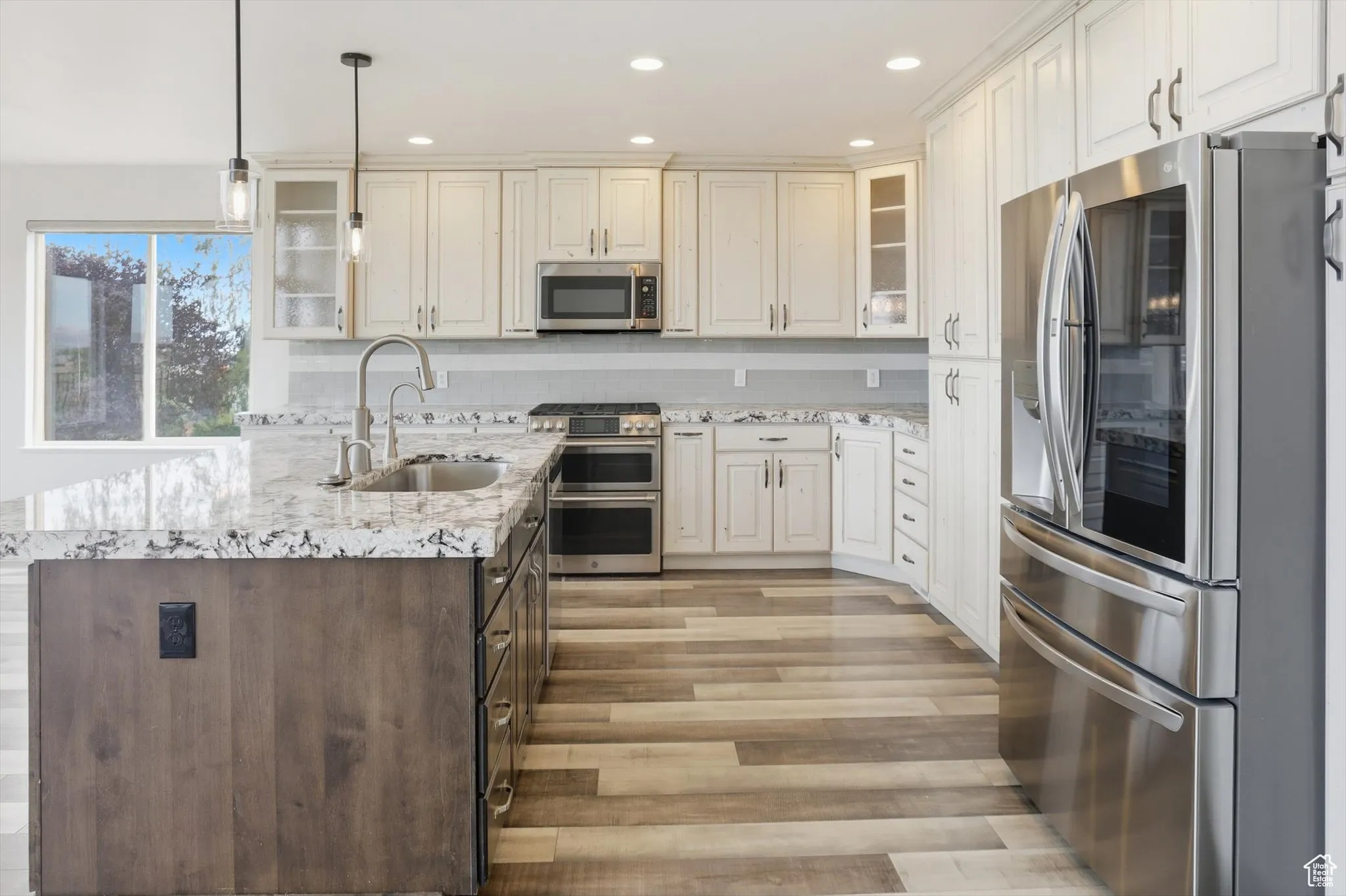 Kitchen with glass insert cabinets, stainless steel appliances, a kitchen island with sink, light stone counters, and hanging light fixtures