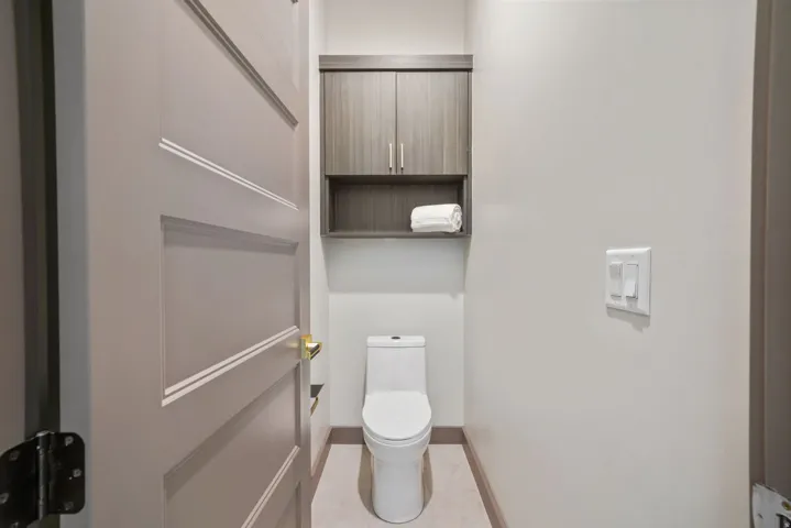 Compact lavatory featuring a white toilet, contemporary wall-mounted cabinetry with an open shelf, light wall paint, and coordinating floor tiles
