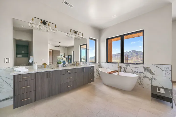 Spacious bathroom featuring a free-standing soaking tub, large windows with mountain scenery, dual vanity with extensive countertop space, full-width mirror, and large format floor tiling