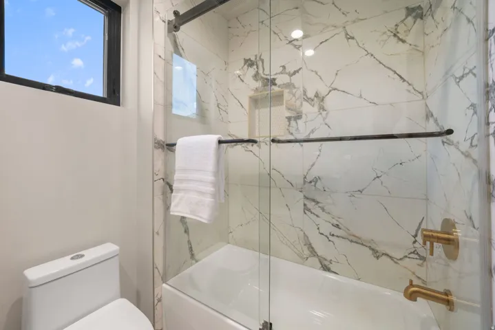Bathroom featuring a tub and shower combination with glass enclosure, large format marble-look tile surround, a built-in shower niche, and brass fixtures
