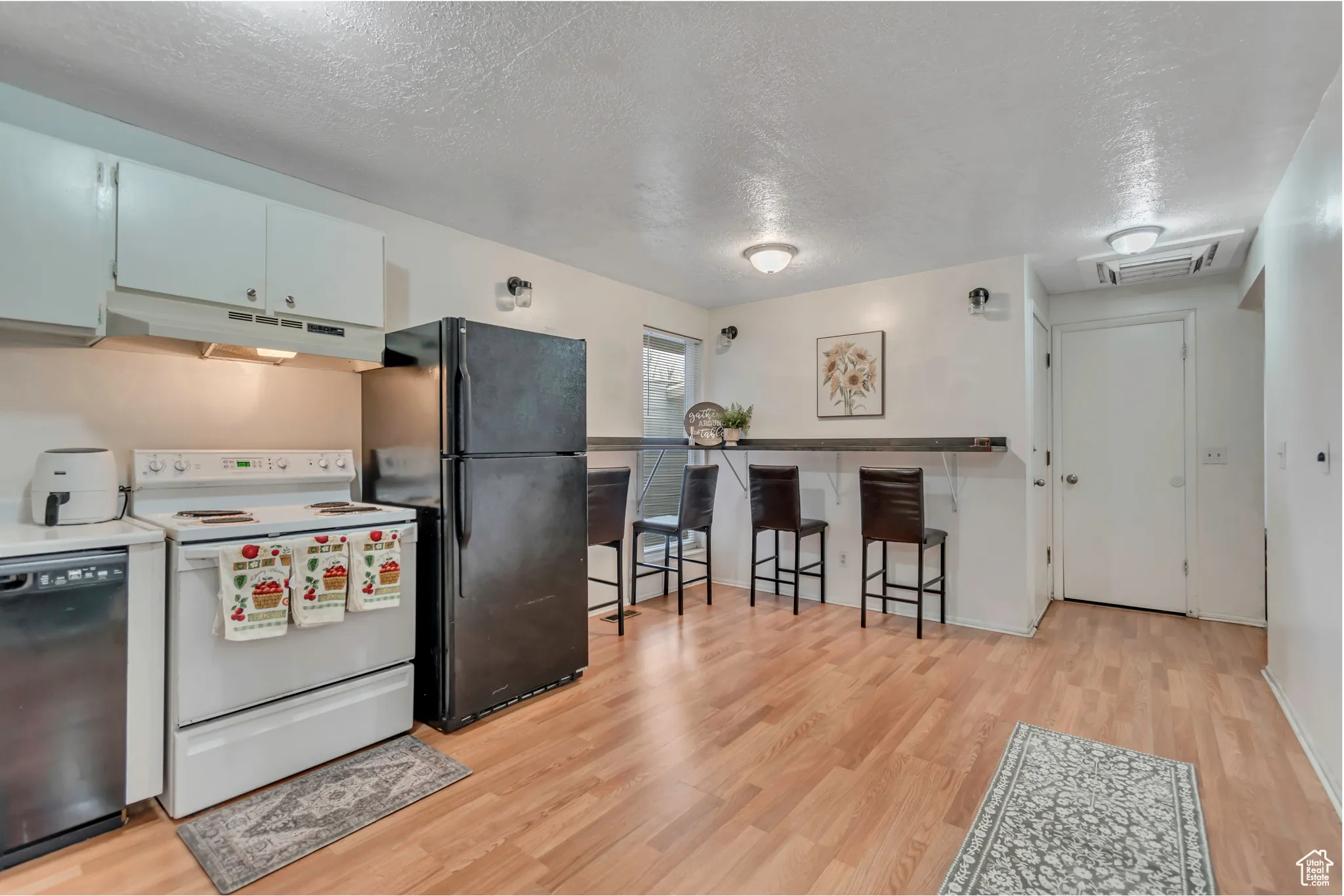 Kitchen featuring black appliances, a textured ceiling, light wood-type flooring, under cabinet range hood, and a kitchen breakfast bar