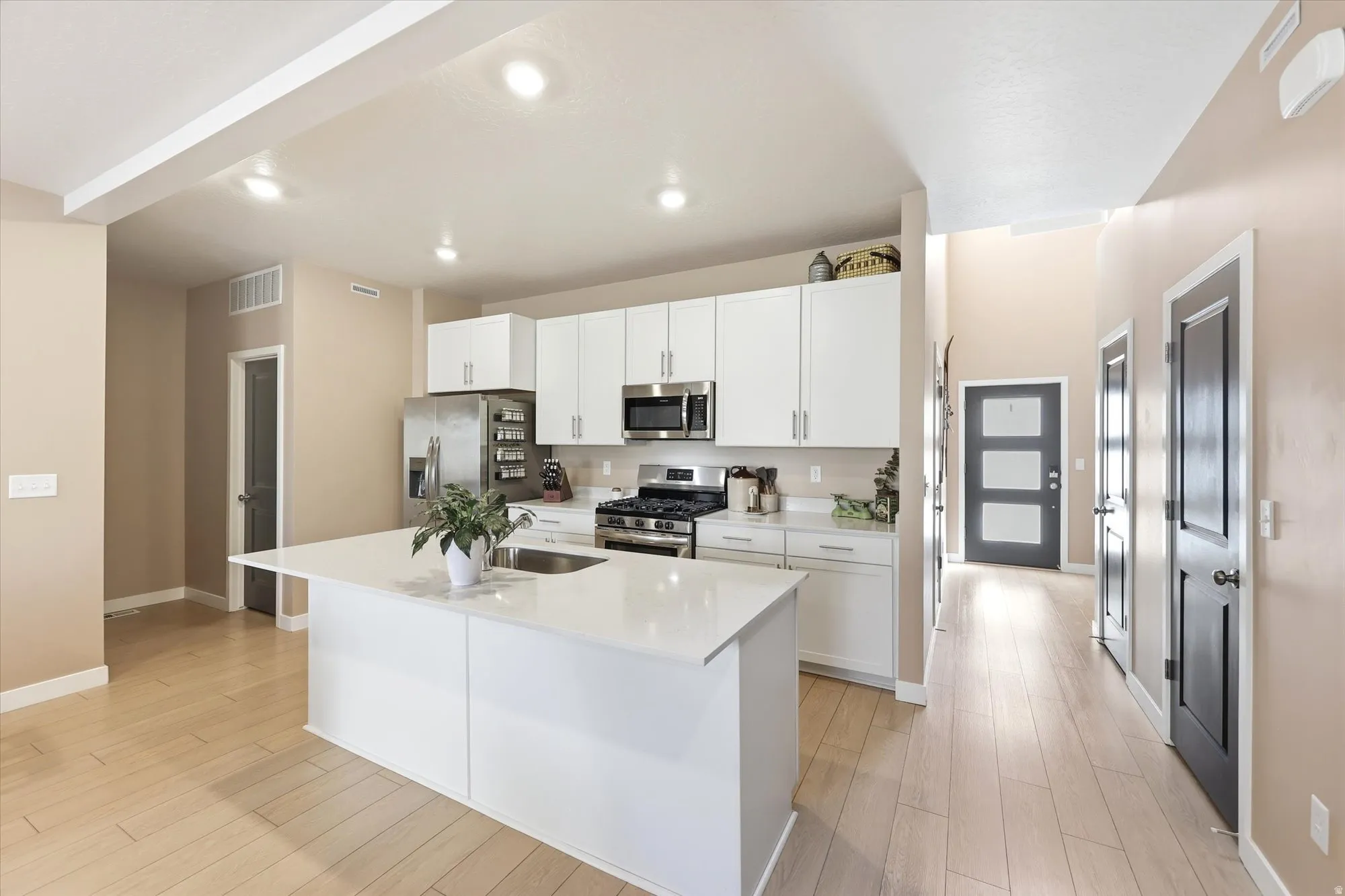 Open-concept kitchen featuring an oversized island with a built-in sink, white shaker cabinetry, stainless steel appliances, and light wood-finish flooring