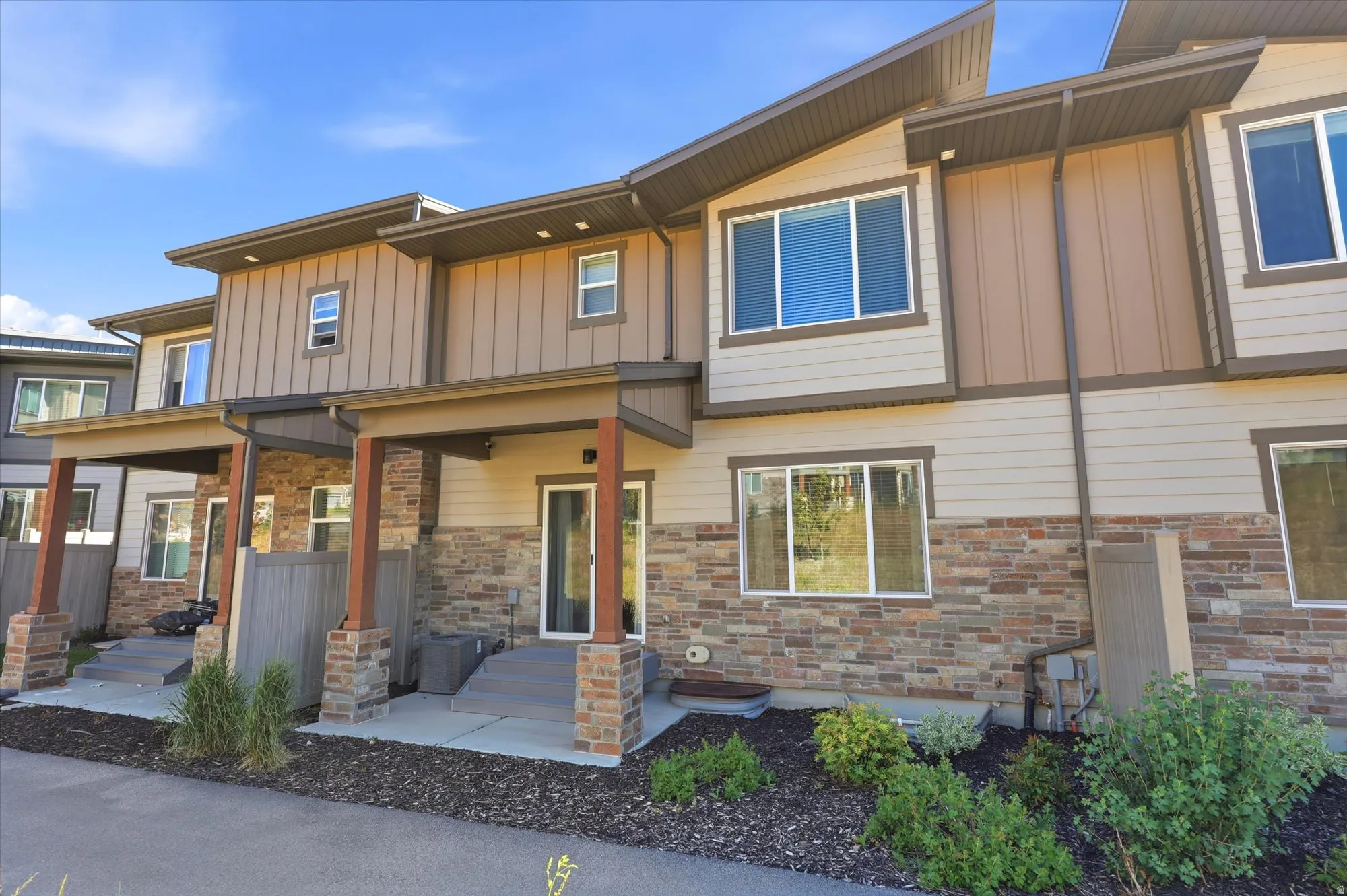 Modern townhome exterior featuring an angular roofline, stone veneer facade, covered patios with wood-look posts, and mature landscaping