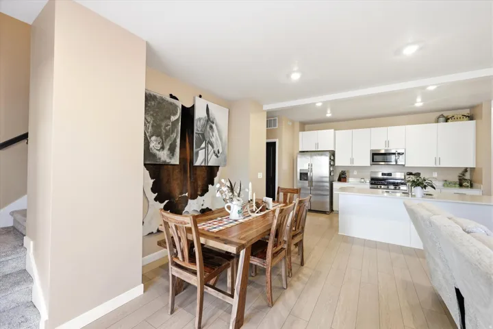 Open-concept living area featuring light wood-finish flooring, recessed lighting, a modern kitchen with white cabinetry and stainless steel appliances, and a staircase with carpeted treads and a dark wood handrail