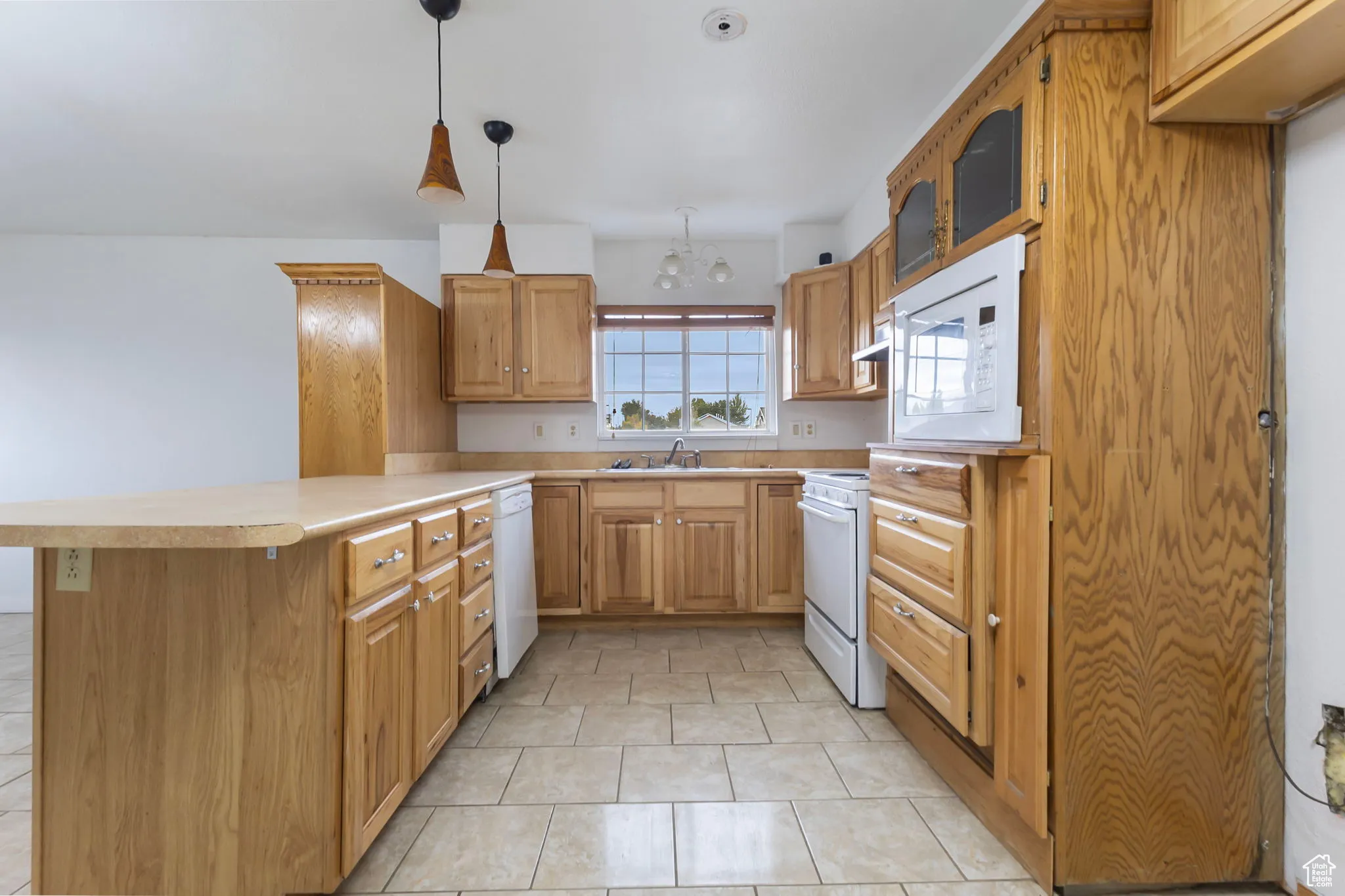 Kitchen with a peninsula, light countertops, light tile patterned flooring, white appliances, and brown cabinetry