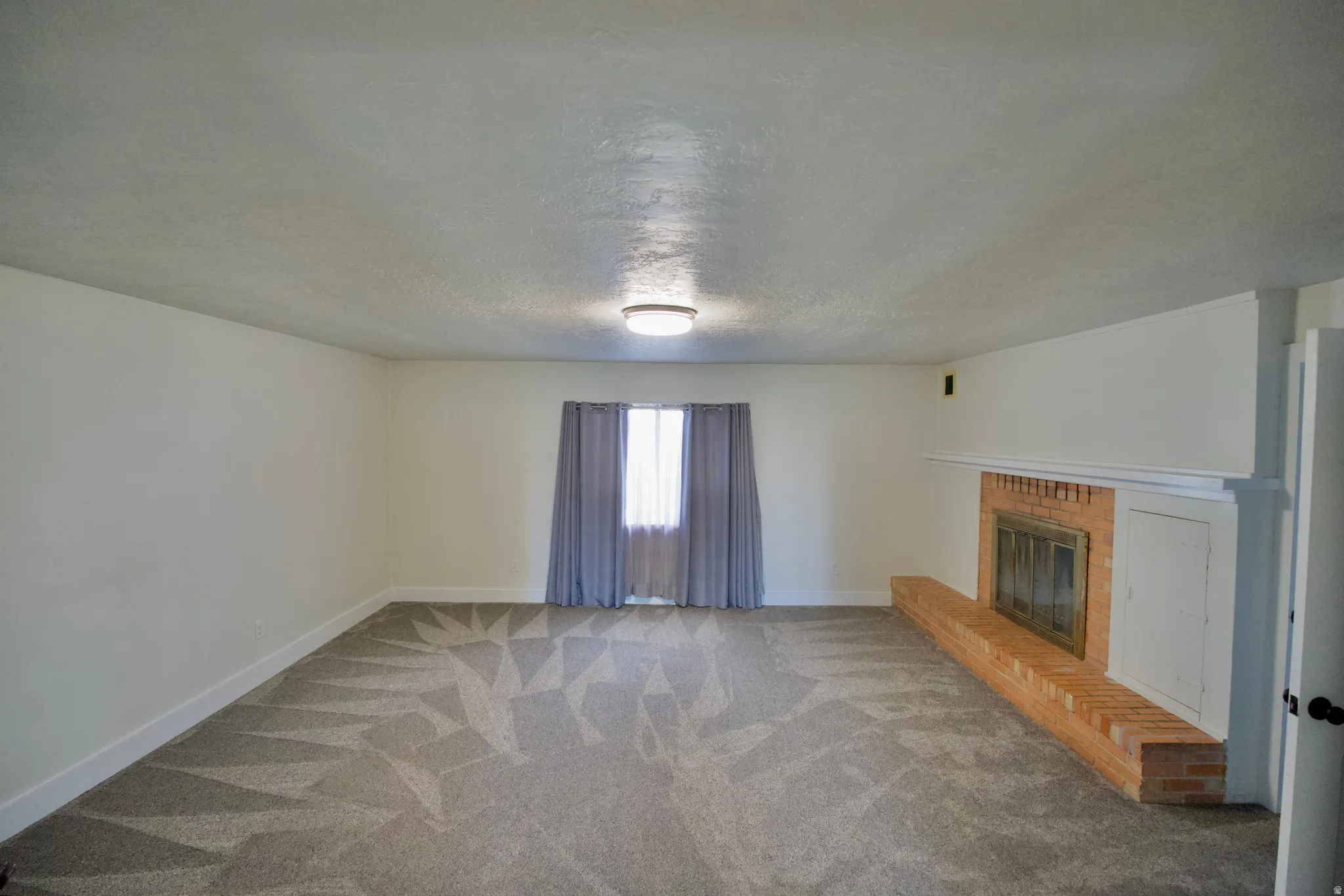 Spacious room featuring patterned carpeting, a traditional brick fireplace with a white mantel, and a window with neutral-toned curtains