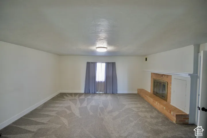 Spacious room featuring patterned carpeting, a traditional brick fireplace with a white mantel, and a window with neutral-toned curtains