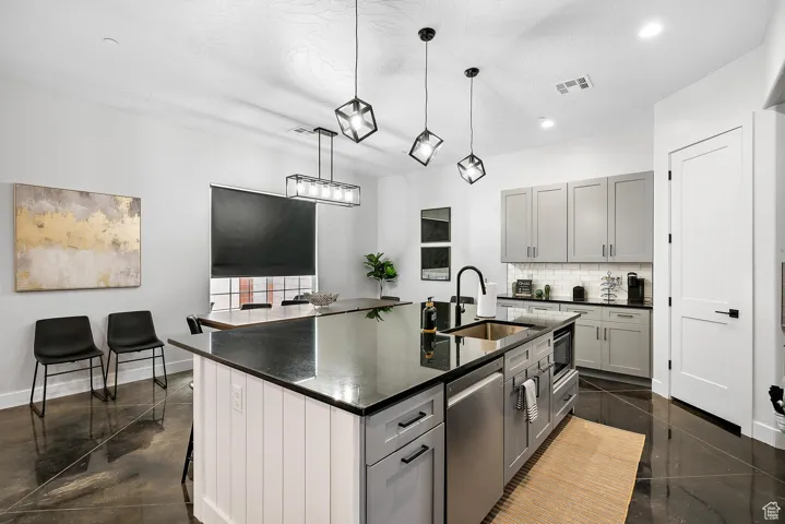 Kitchen with gray cabinets, a center island with sink, tasteful backsplash, hanging light fixtures, and stainless steel dishwasher