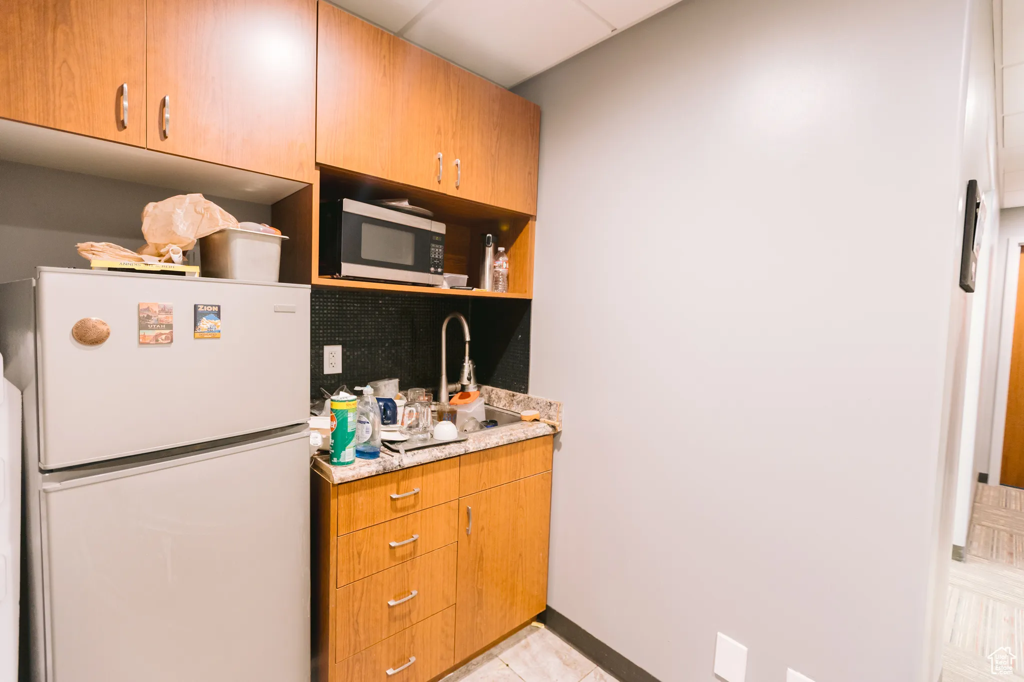 Kitchen featuring freestanding refrigerator, light tile patterned floors, stainless steel microwave, open shelves, and brown cabinets