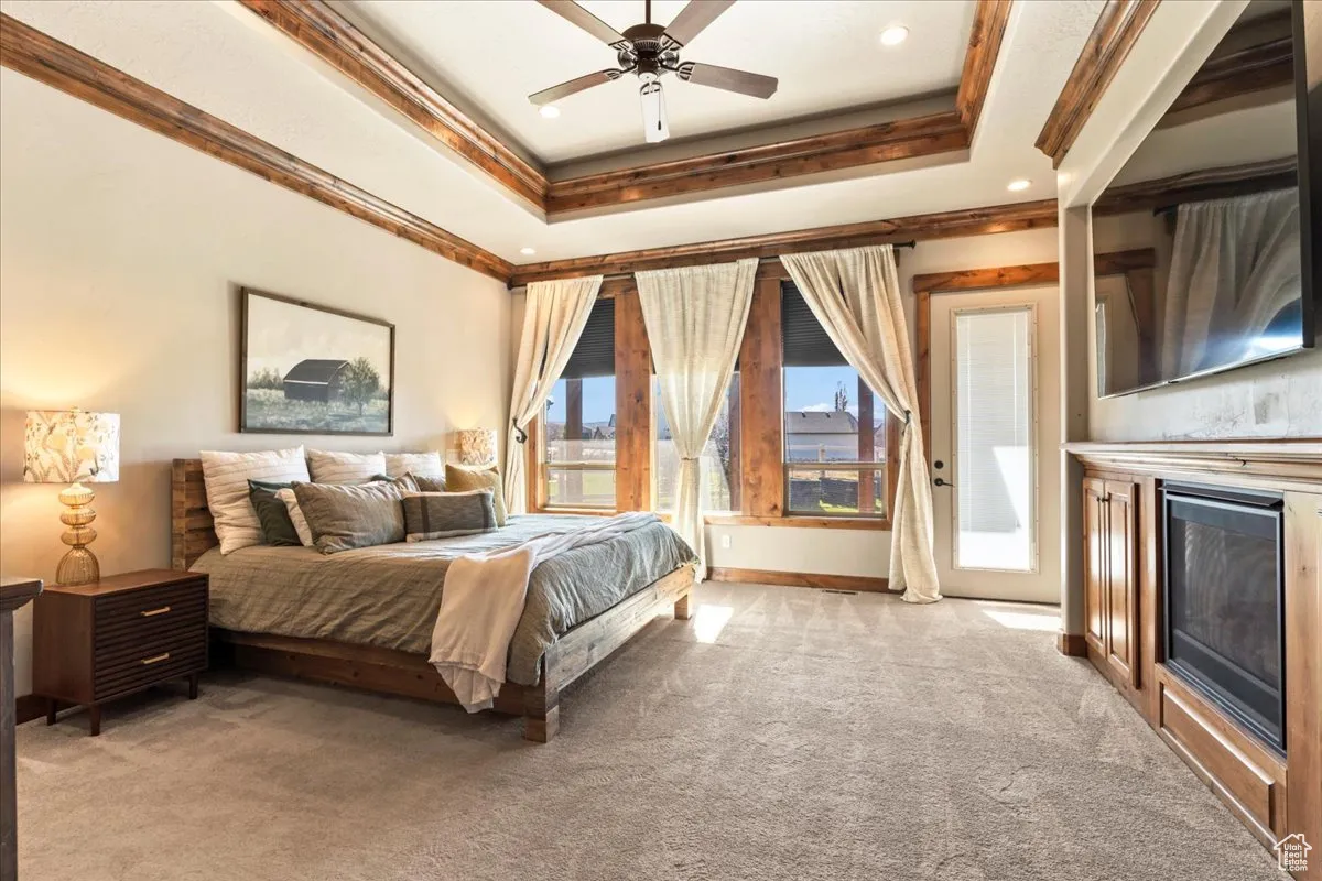 Primary Bedroom with light carpet, a raised ceiling, a glass covered fireplace, ornamental molding, and ceiling fan