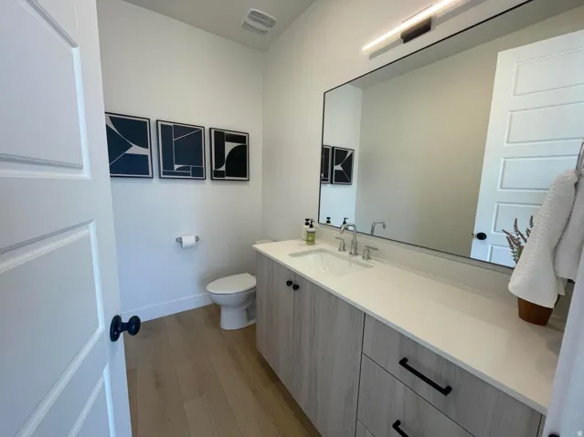 Wood-finish flooring with a toilet and a vanity featuring a white countertop, built-in sink, and wood-finish cabinetry with black hardware