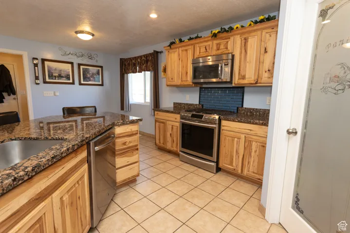 Kitchen featuring appliances with stainless steel finishes, dark stone counters, light tile patterned floors, a textured ceiling, and backsplash