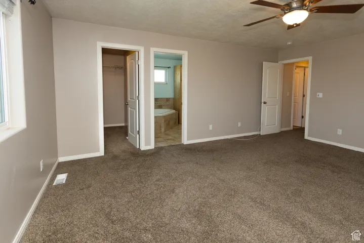 Unfurnished bedroom with a textured ceiling, carpet, a spacious closet, ceiling fan, and ensuite bathroom