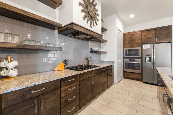 Kitchen with open shelves, dark brown cabinetry, tasteful backsplash, stainless steel appliances, and light stone counters