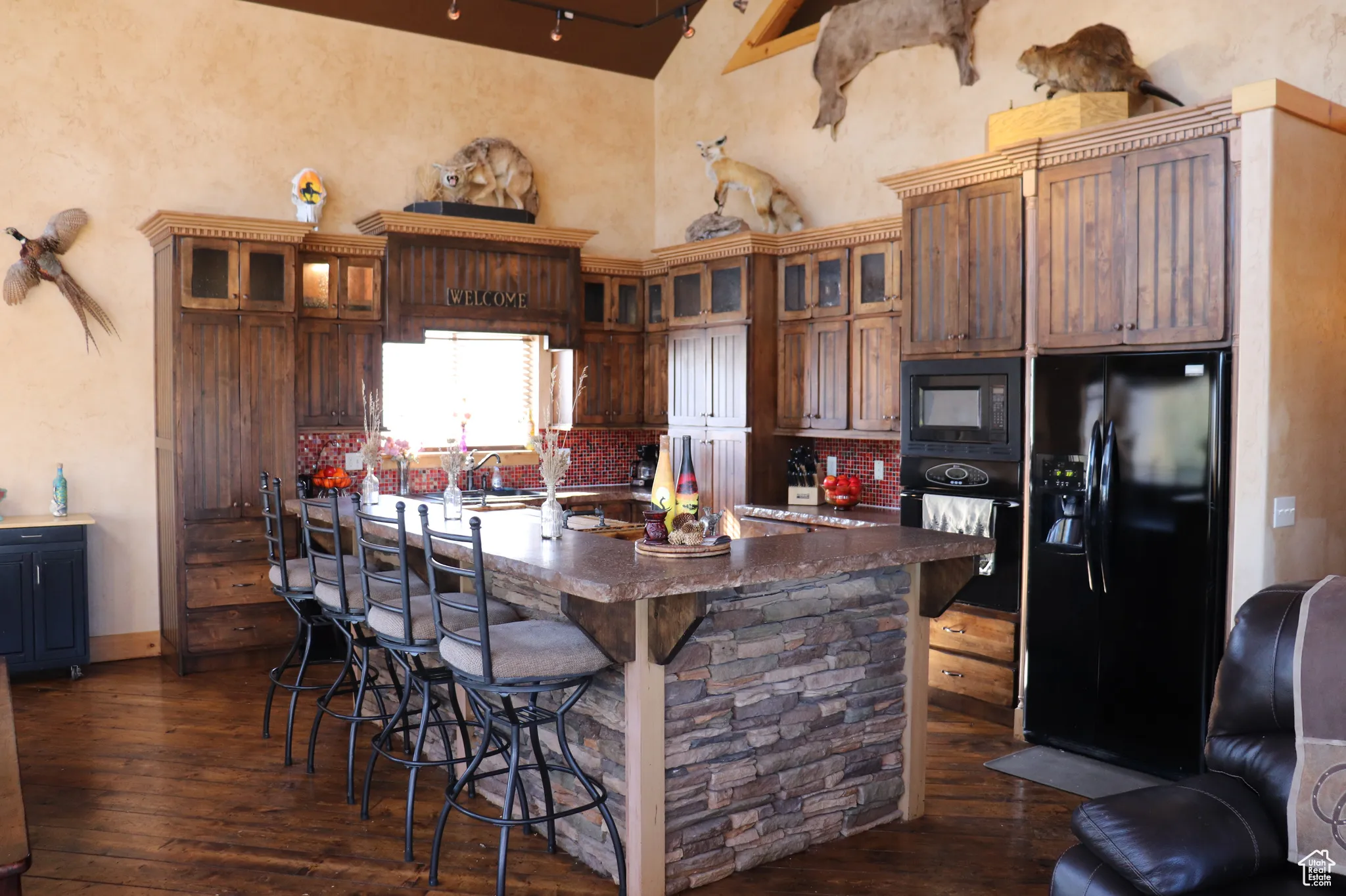 Kitchen with black appliances, a breakfast bar area, dark wood-style floors, a large island with sink, and glass insert cabinets