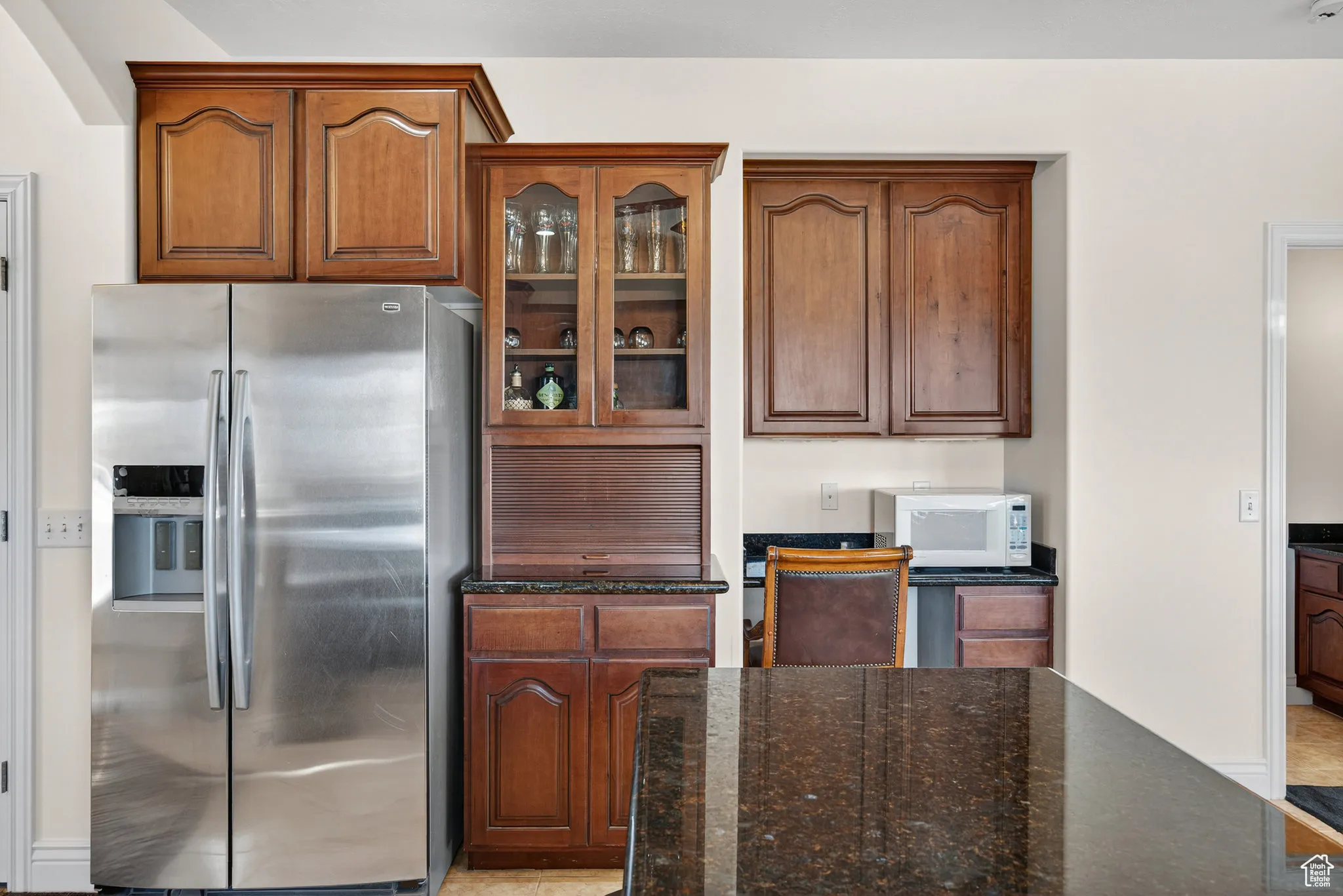 Kitchen featuring stainless steel refrigerator with ice dispenser, dark stone counters, glass insert cabinets.