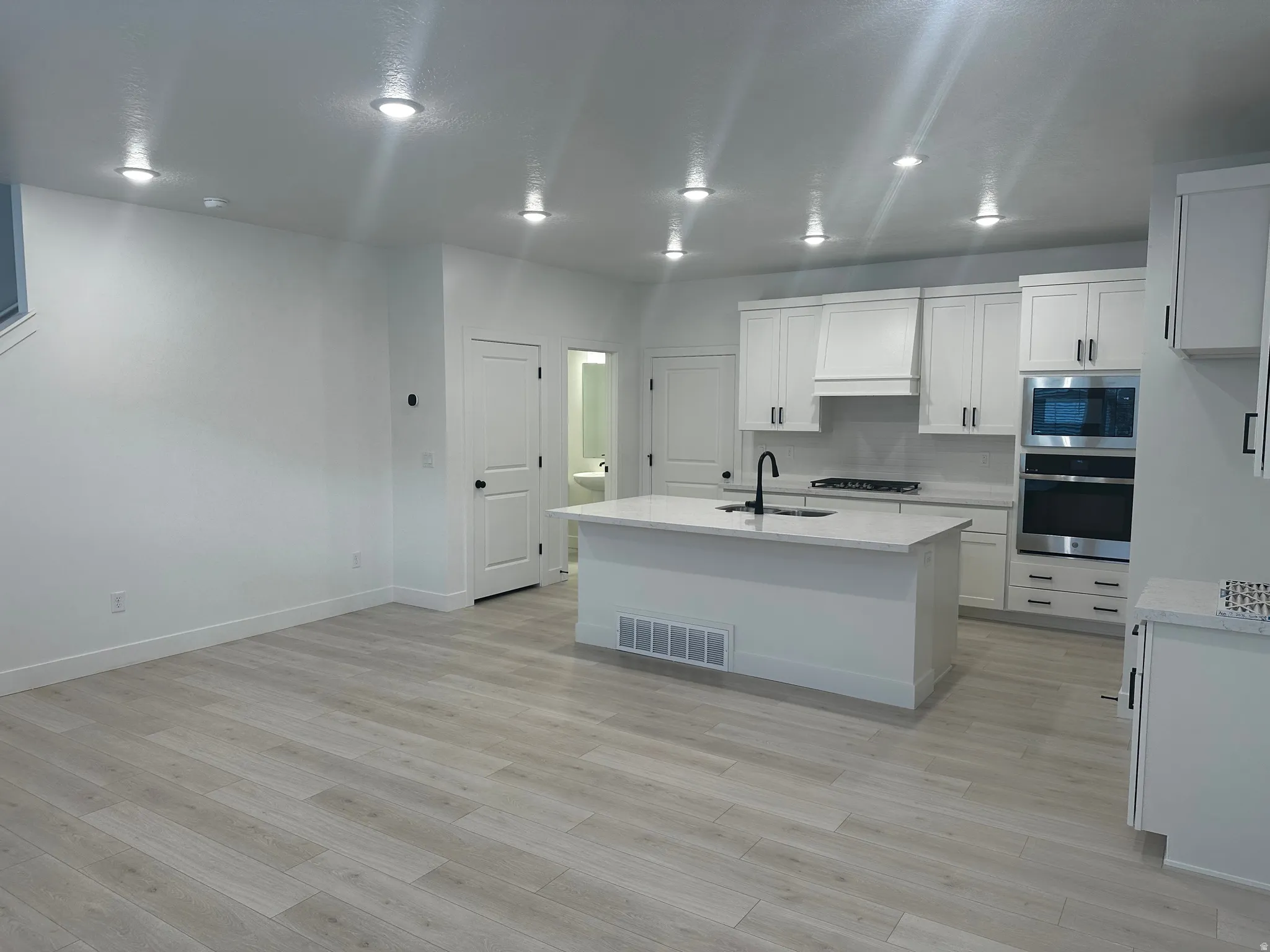 Kitchen with a kitchen island with sink, stainless steel appliances, white cabinets, light stone countertops, and light wood finished floors