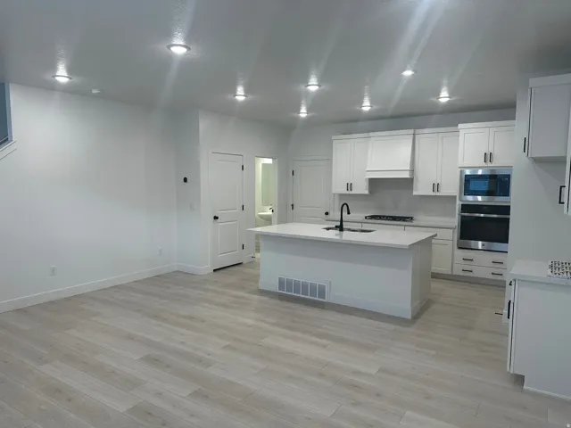 Kitchen with a kitchen island with sink, stainless steel appliances, white cabinets, light stone countertops, and light wood finished floors