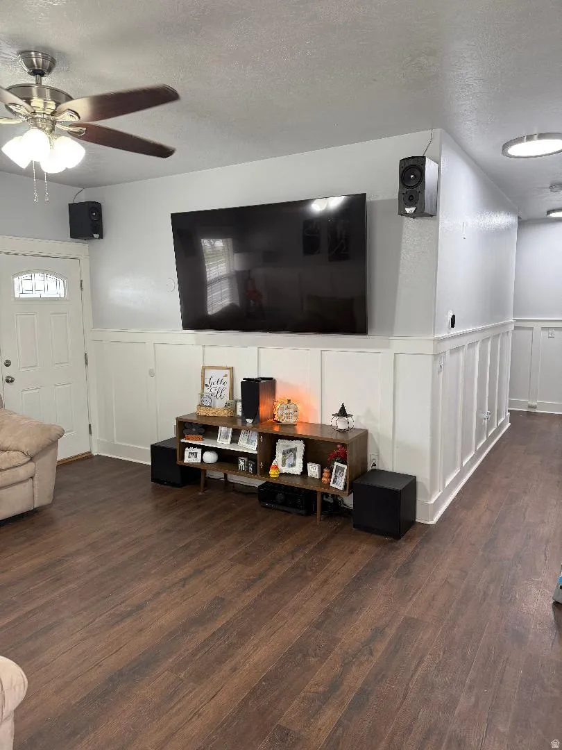 Living area featuring a decorative wall, a textured ceiling, wainscoting, dark wood-style floors, and ceiling fan