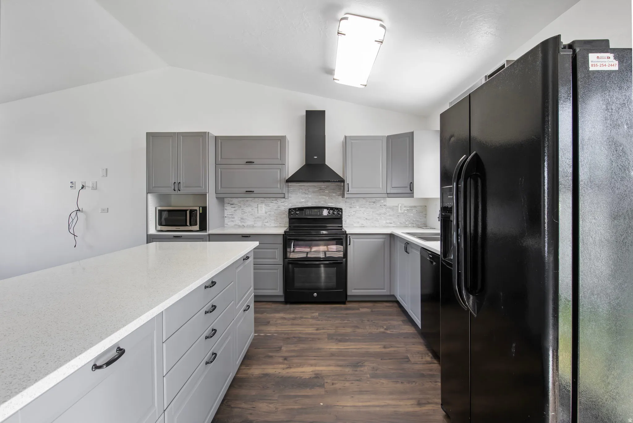 Kitchen featuring gray cabinetry, black appliances, vaulted ceiling, wall chimney exhaust hood, and dark wood-style flooring