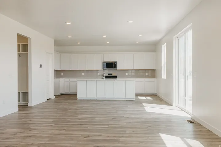 Spacious open-concept living area featuring wood-finish flooring, white cabinetry, a kitchen island, integrated stainless steel appliances, and a sliding glass door