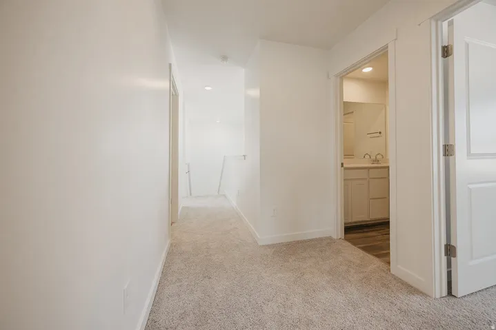 Hallway and ensuite featuring neutral carpet flooring, white painted walls, recessed ceiling lighting, and direct access to a full bathroom with dual vanity and warm wood cabinetry