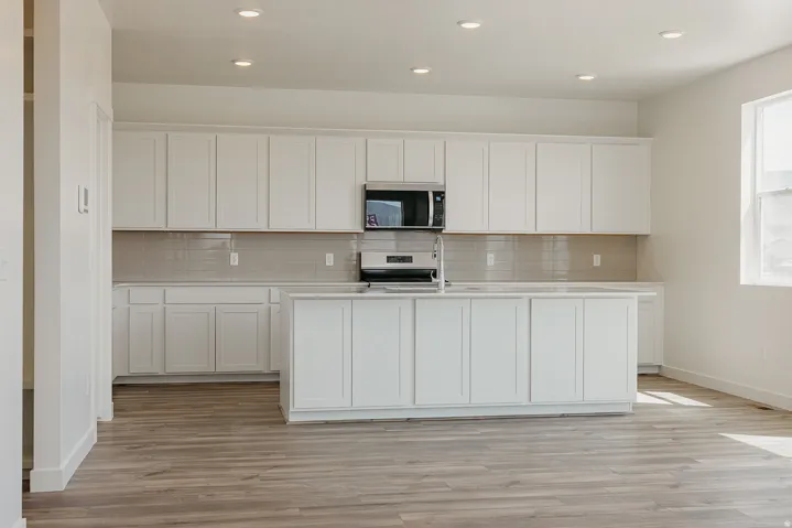 Open-concept kitchen and dining area featuring white shaker-style cabinetry, stainless steel appliances, neutral tile backsplash, a large center island with storage, and wood-finish flooring throughout