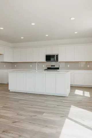 Spacious kitchen featuring white cabinetry, a large island with sink, stainless steel appliances, a light-toned tile backsplash, and wood-finish flooring