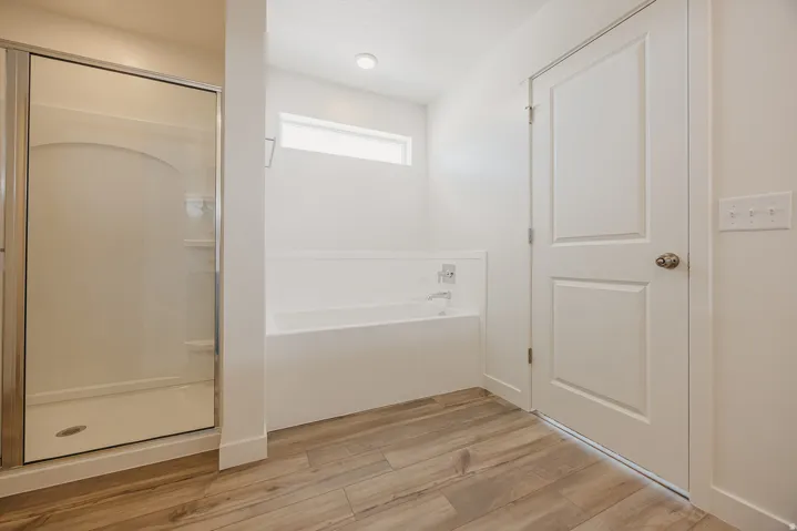 Full shower enclosure with glass door, deep soaking tub, wood-finish flooring, and white paneled door