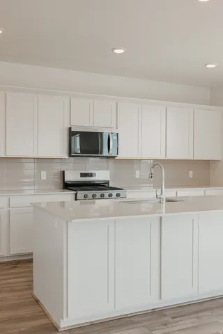 White shaker cabinetry, stainless steel appliances, light tone wood-finish flooring, recessed lighting, and a kitchen island with sink