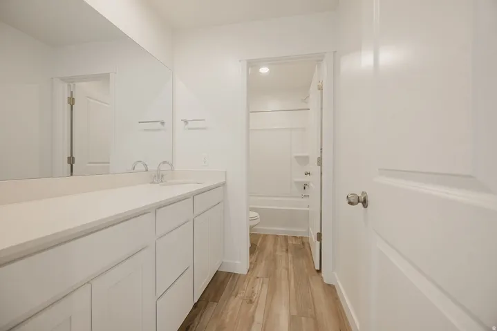 White vanity with integrated sink, expansive mirror, wood-finish flooring, and a separate tub/shower combination