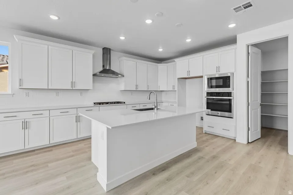 Kitchen with appliances with stainless steel finishes, wall chimney exhaust hood, light wood-type flooring, recessed lighting, and an island with sink