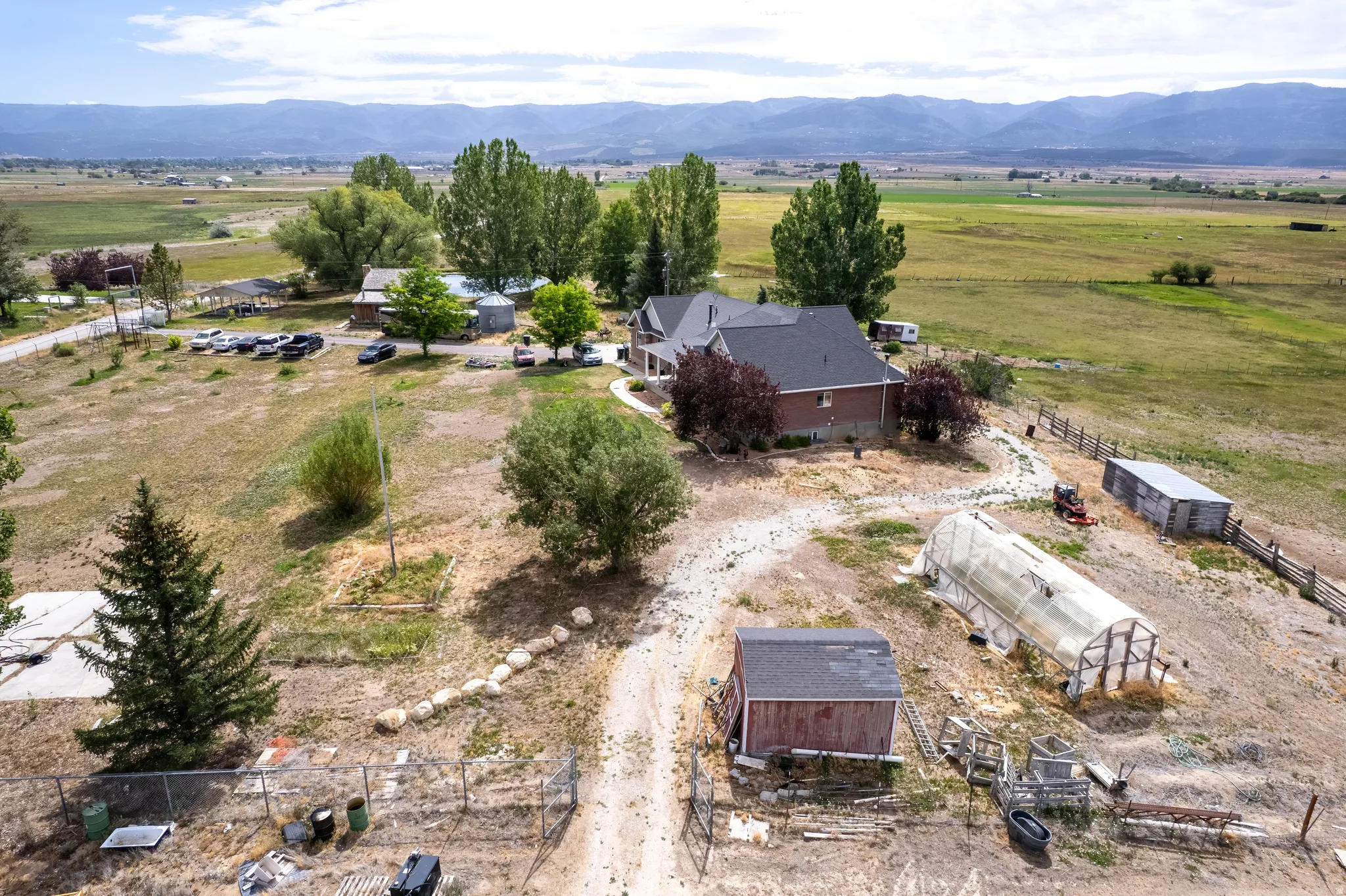 Expansive property featuring a brick residence with a dark roof, multiple outbuildings, a greenhouse structure, mature trees, and mountainous terrain in the background