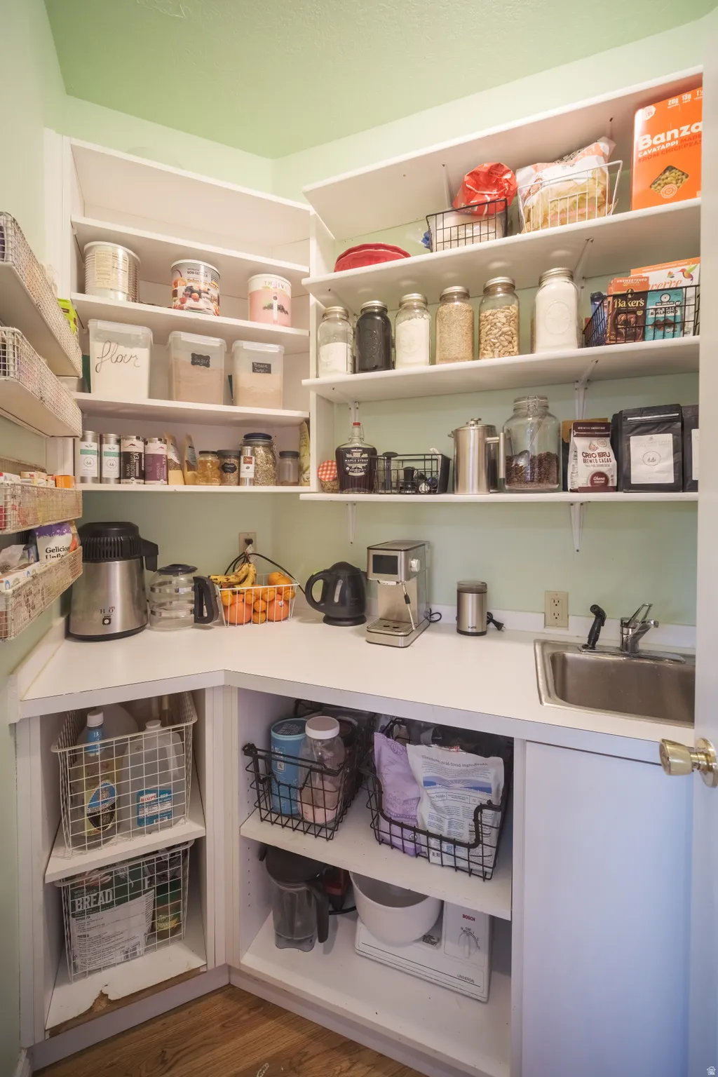 Corner pantry featuring extensive white shelving, a built-in stainless steel sink, wood-finish flooring, white countertops, and integrated lower cabinetry