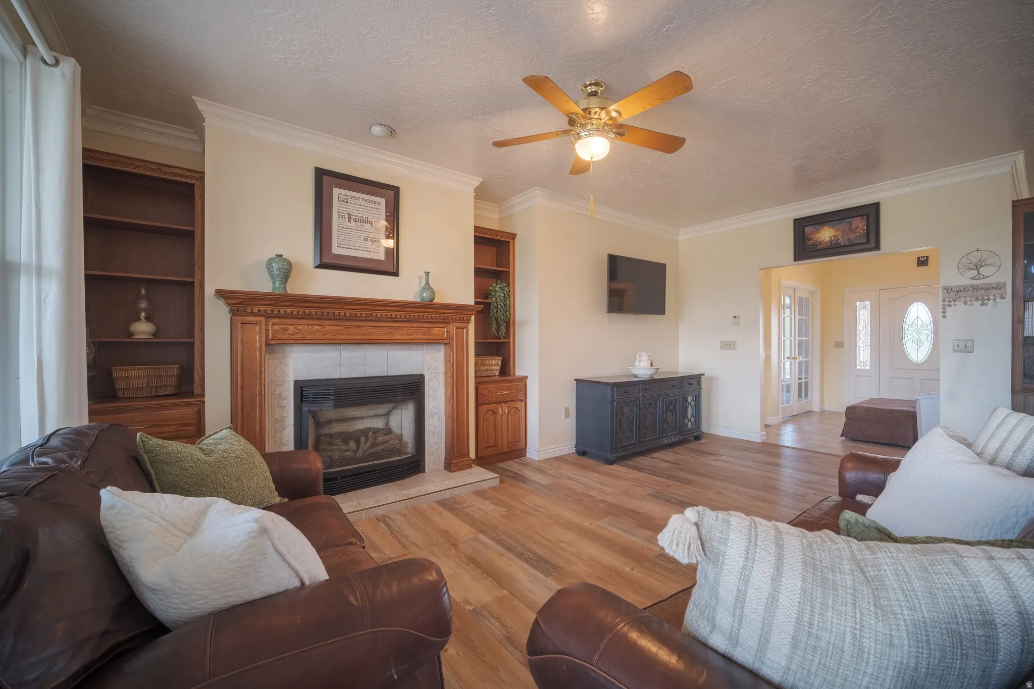 Living area featuring wood-finish flooring, a fireplace with a wood mantle and tile surround, built-in wooden shelving, crown molding, and a ceiling fan