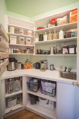 Corner pantry featuring extensive white shelving, a built-in stainless steel sink, wood-finish flooring, white countertops, and integrated lower cabinetry